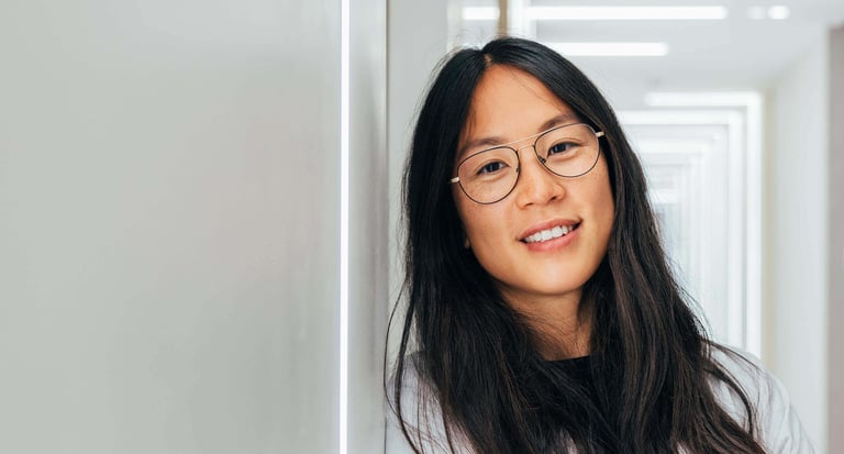 A woman wearing glasses leans casually against a wall, exuding a relaxed and thoughtful demeanor.