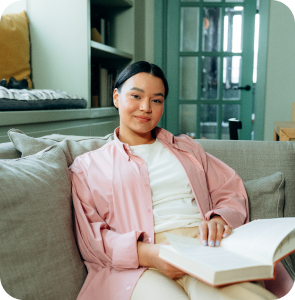 A woman seated on a couch, engrossed in reading a book, surrounded by a cozy living room atmosphere.