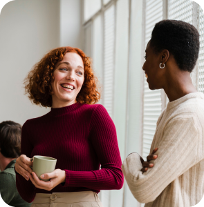 Two women smiling and conversing in an office environment, showcasing a friendly and collaborative atmosphere.